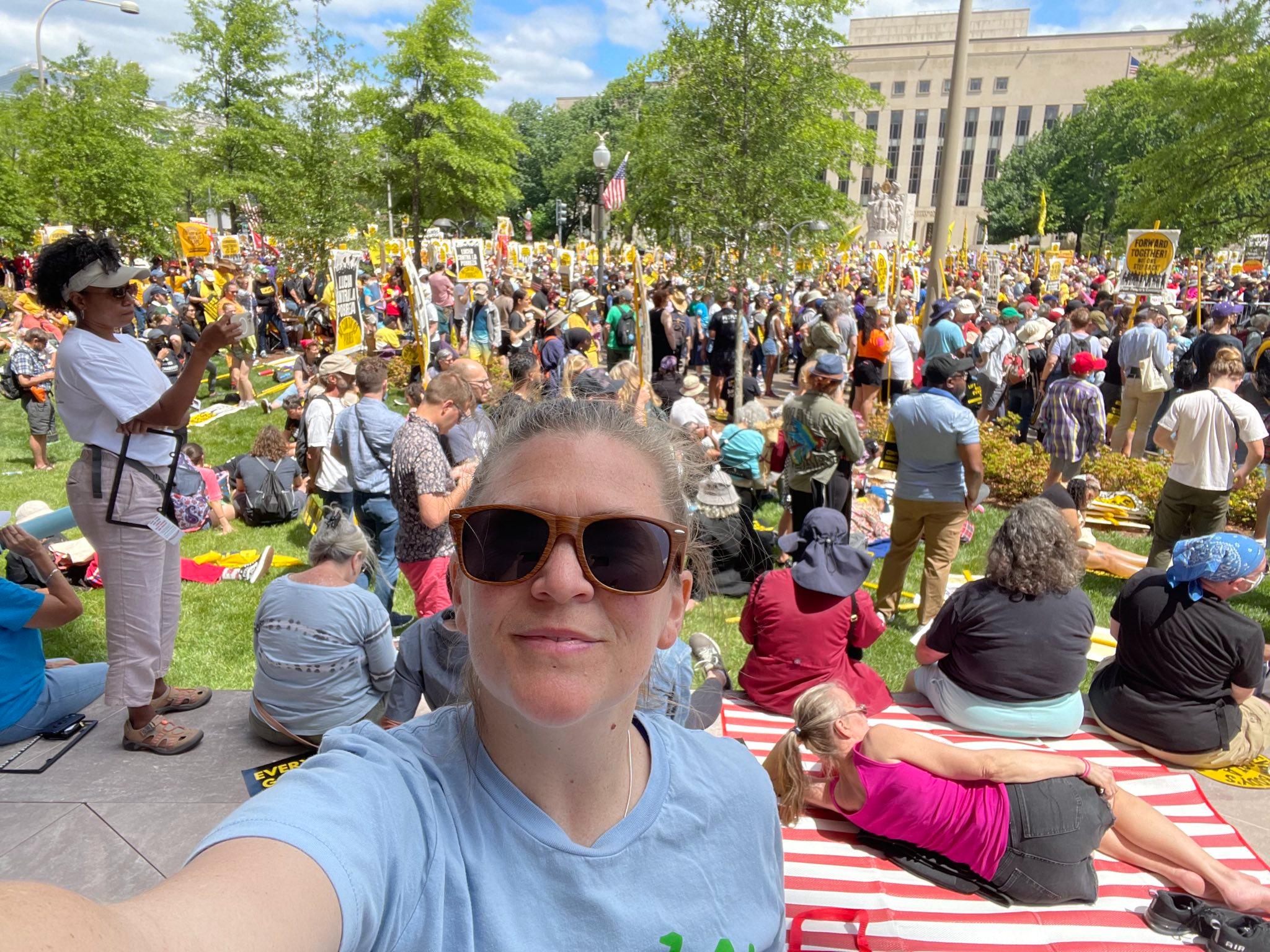 A person with sunglasses at a political rally. In the background: people holding up signs